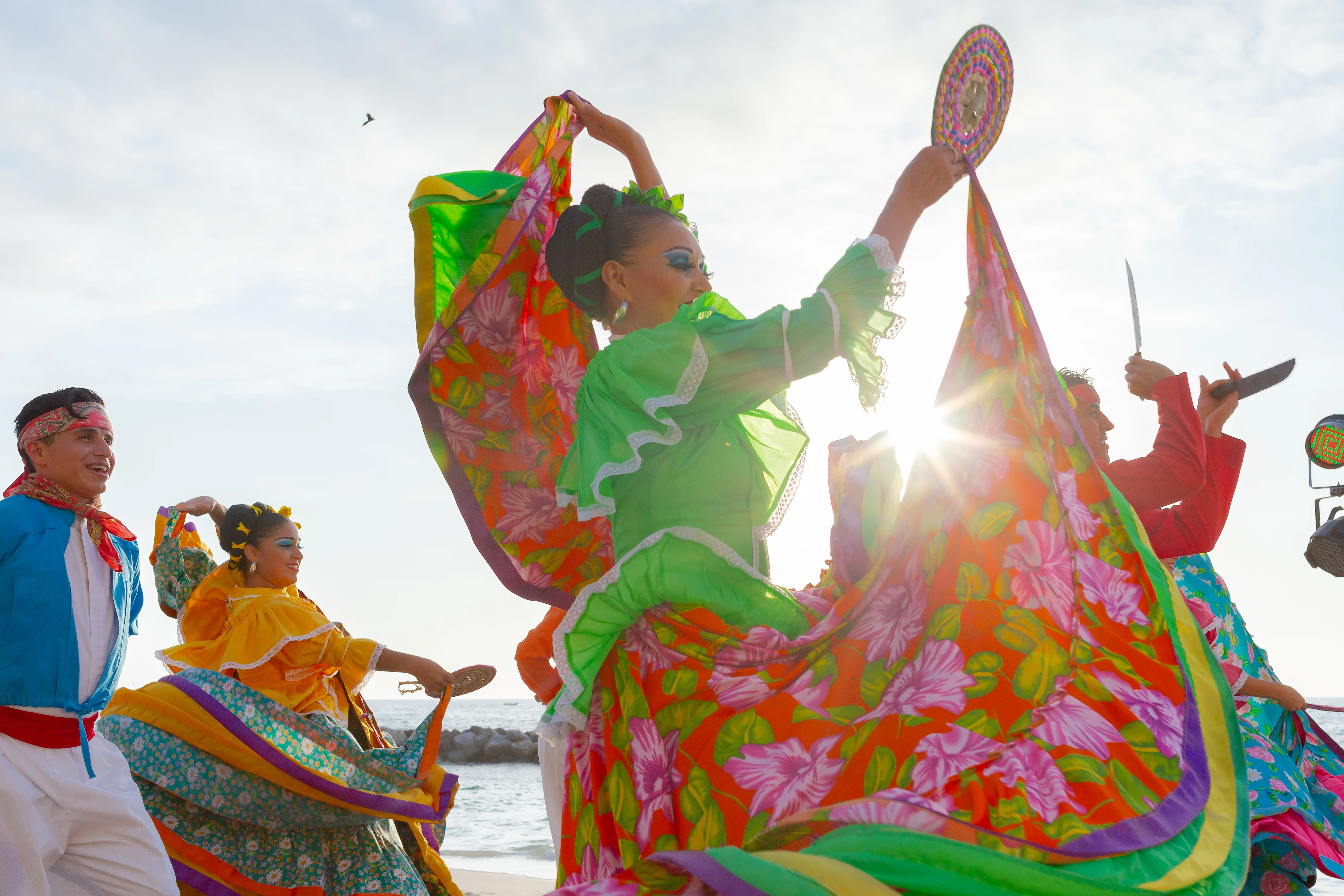 Dancers in vibrant traditional Mexican attire perform a lively dance on the beach at sunset.