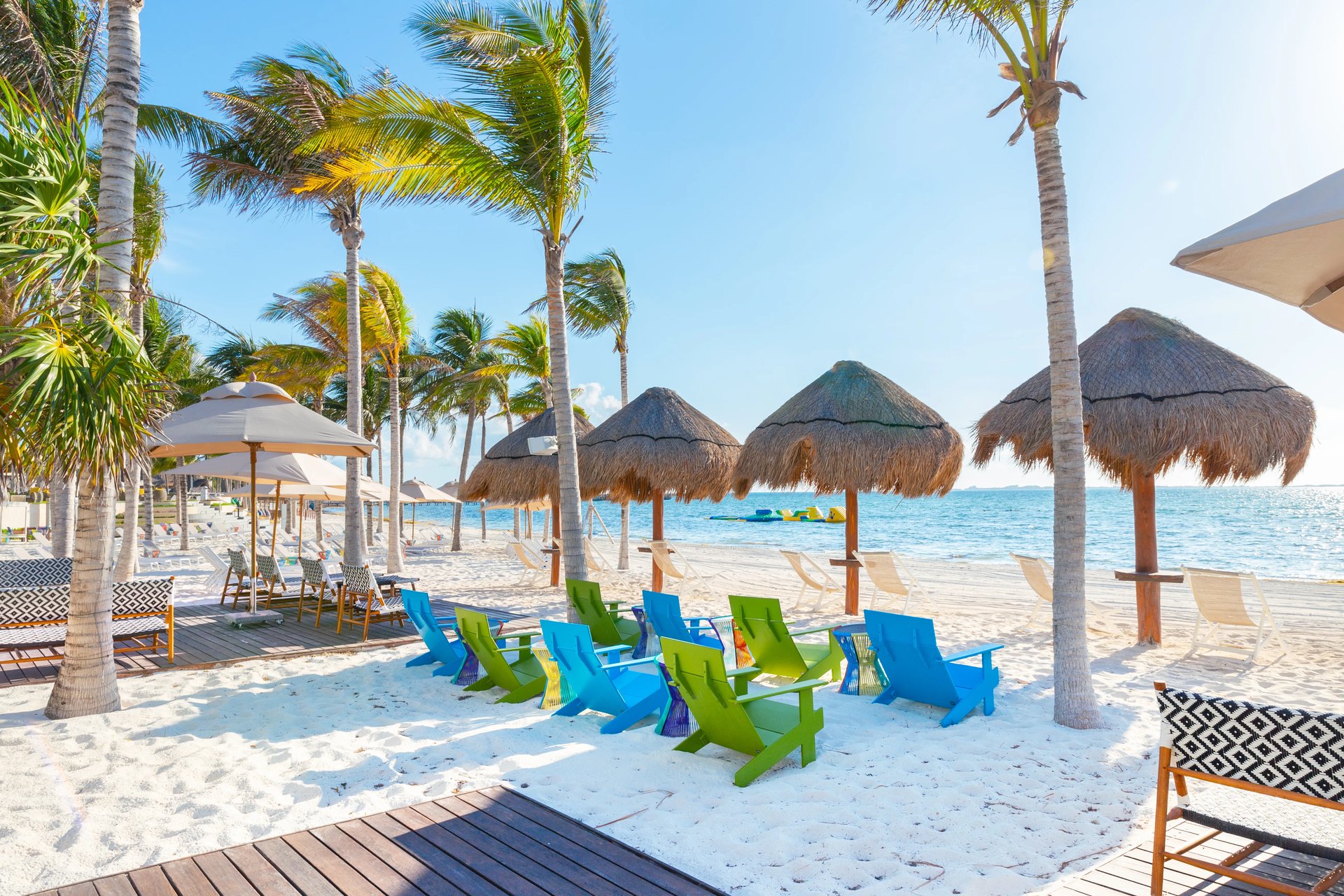 Sunlit Garza Blanca beachfront in Mexico with swaying palms, thatch palapas, and colorful blue and green Adirondack chairs facing turquoise sea.