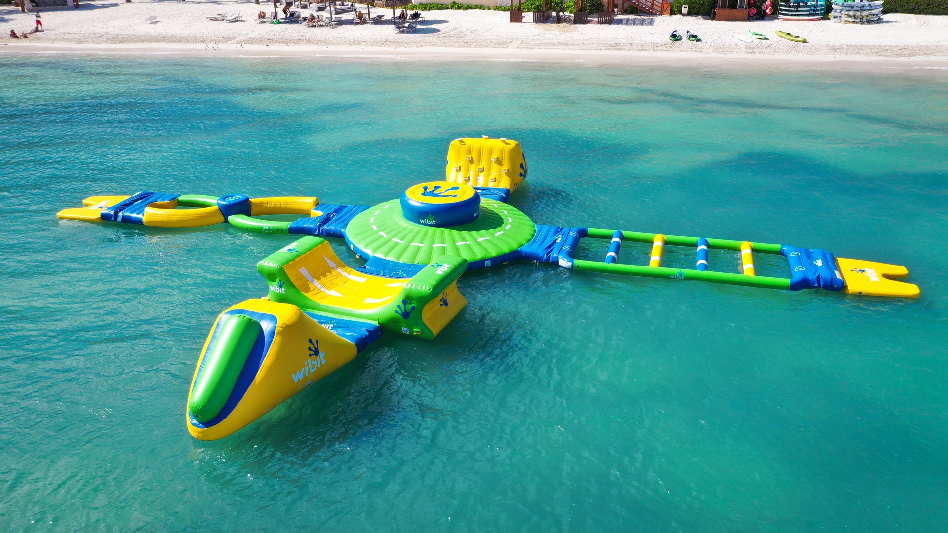 Colorful inflatable water obstacle course floats on crystal turquoise sea beside a white-sand beach.