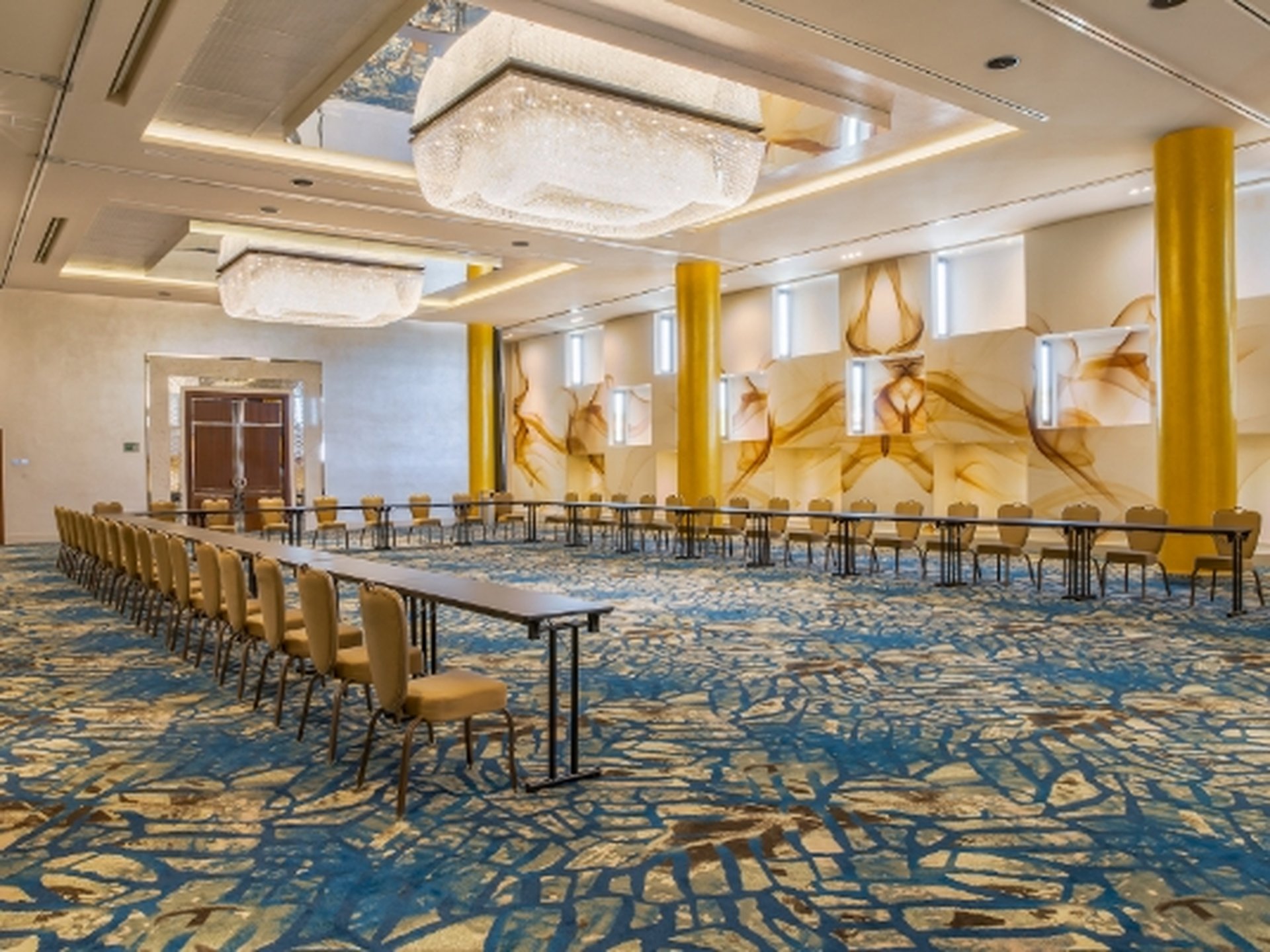 Spacious conference room with U-shaped table setup, elegant chandeliers, abstract wall art, and blue patterned carpet.