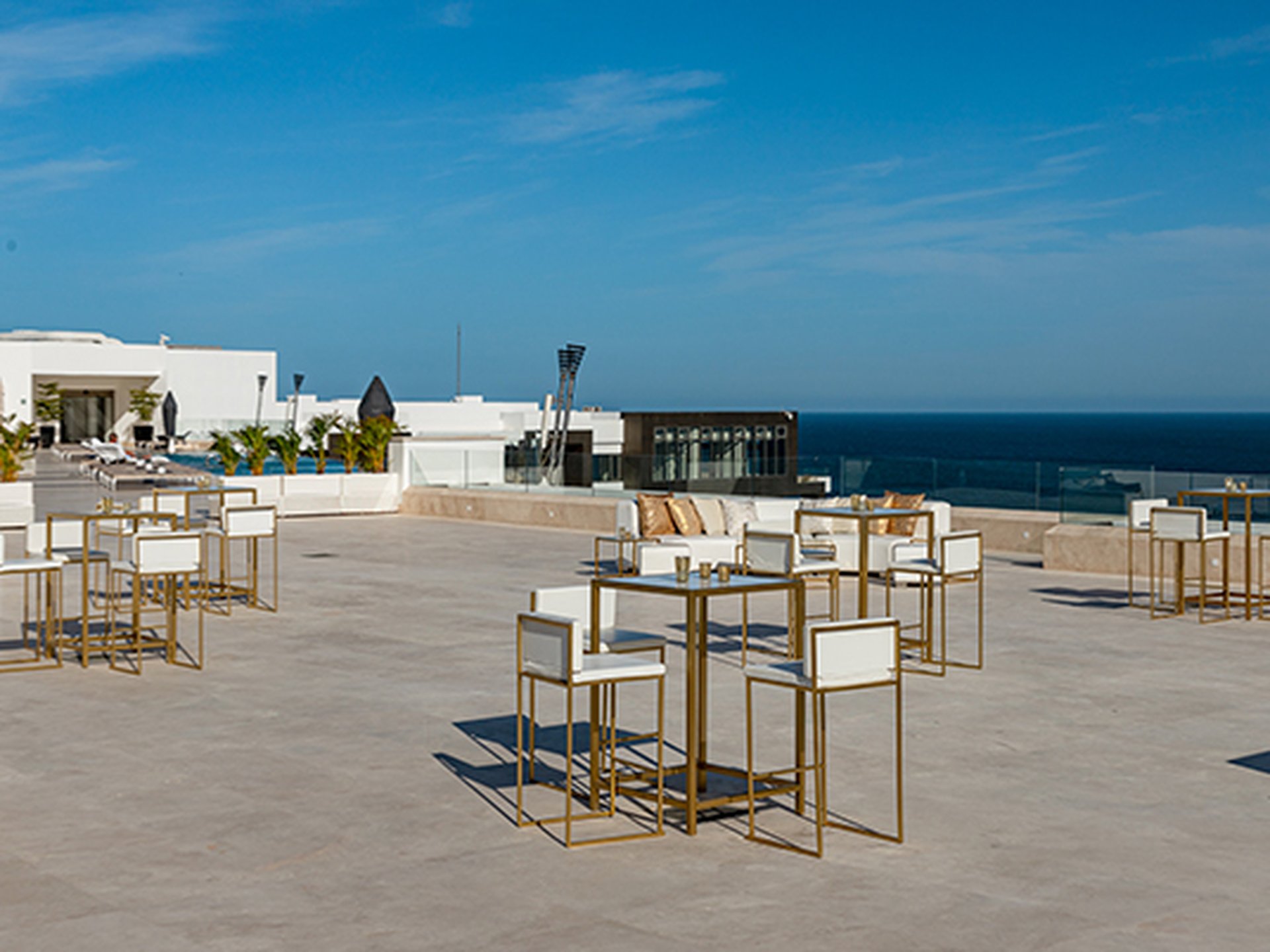 Rooftop terrace with white and gold tables and chairs, overlooking the ocean. Clear blue sky and modern architecture in the background.