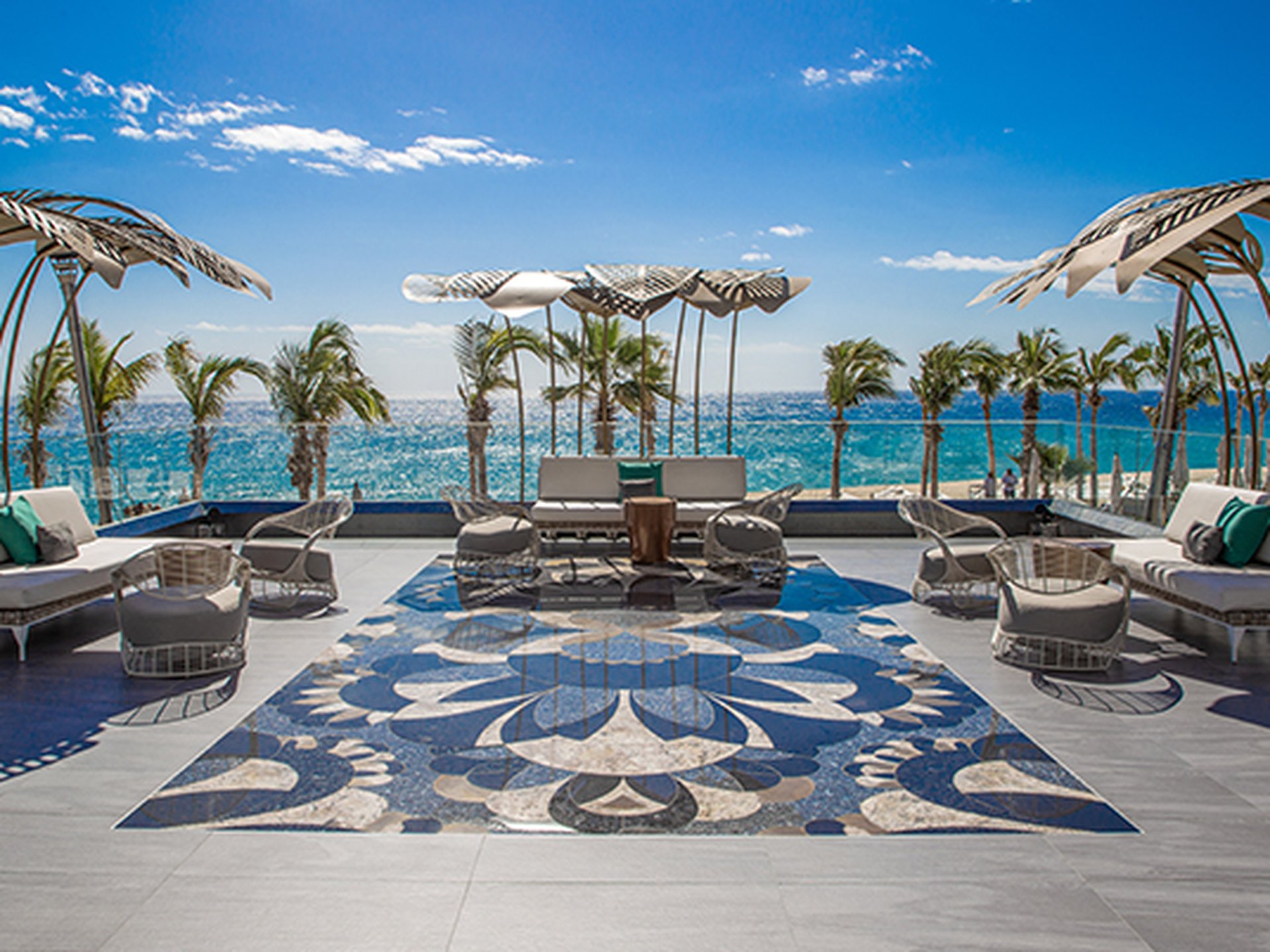 Luxurious oceanfront patio with stylish lounge chairs, artistic mosaic floor, and palm trees under a clear blue sky.