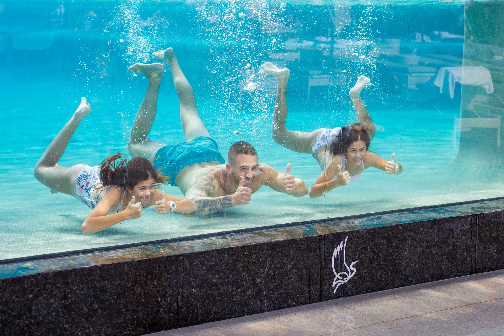 Underwater family fun: four guests, two kids and two adults, smile and thumbs-up in a turquoise Garza Blanca resort pool, México.