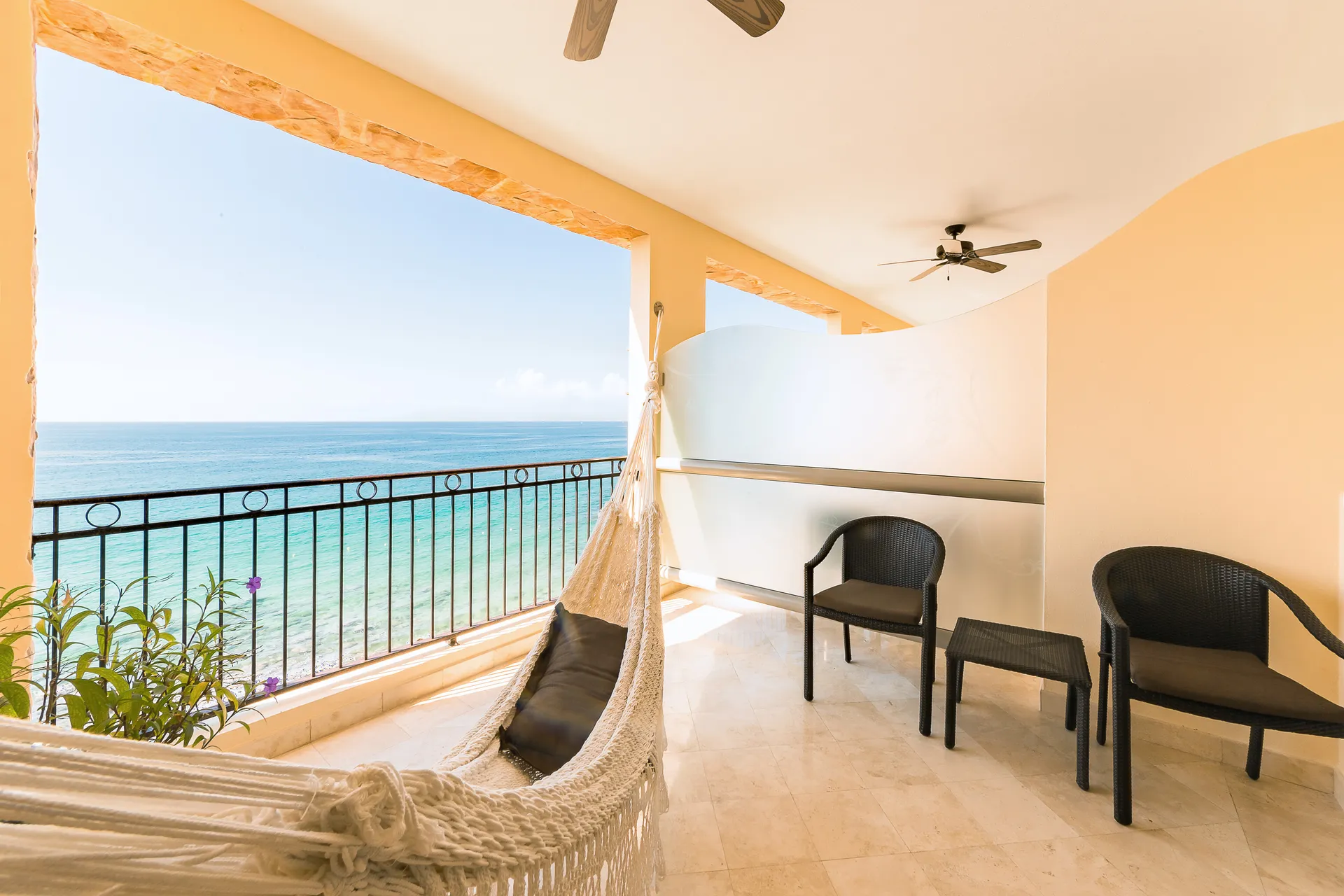 Private balcony with a hammock, two chairs, and ocean view at a luxury resort. Bright, serene setting with blue skies and calm sea.