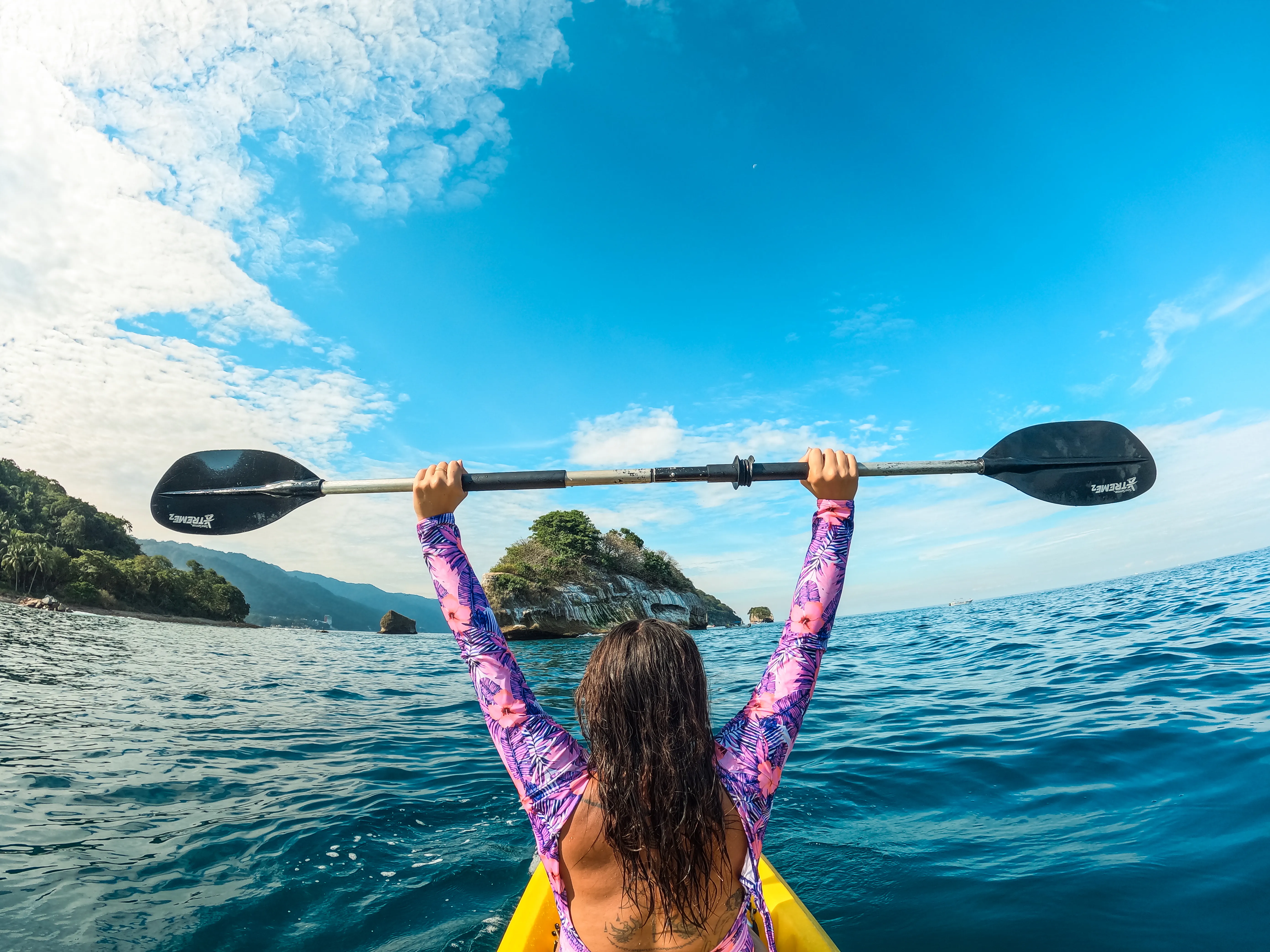 Person in a kayak holds a paddle triumphantly above their head on a sunny, blue ocean with distant rocky islands.