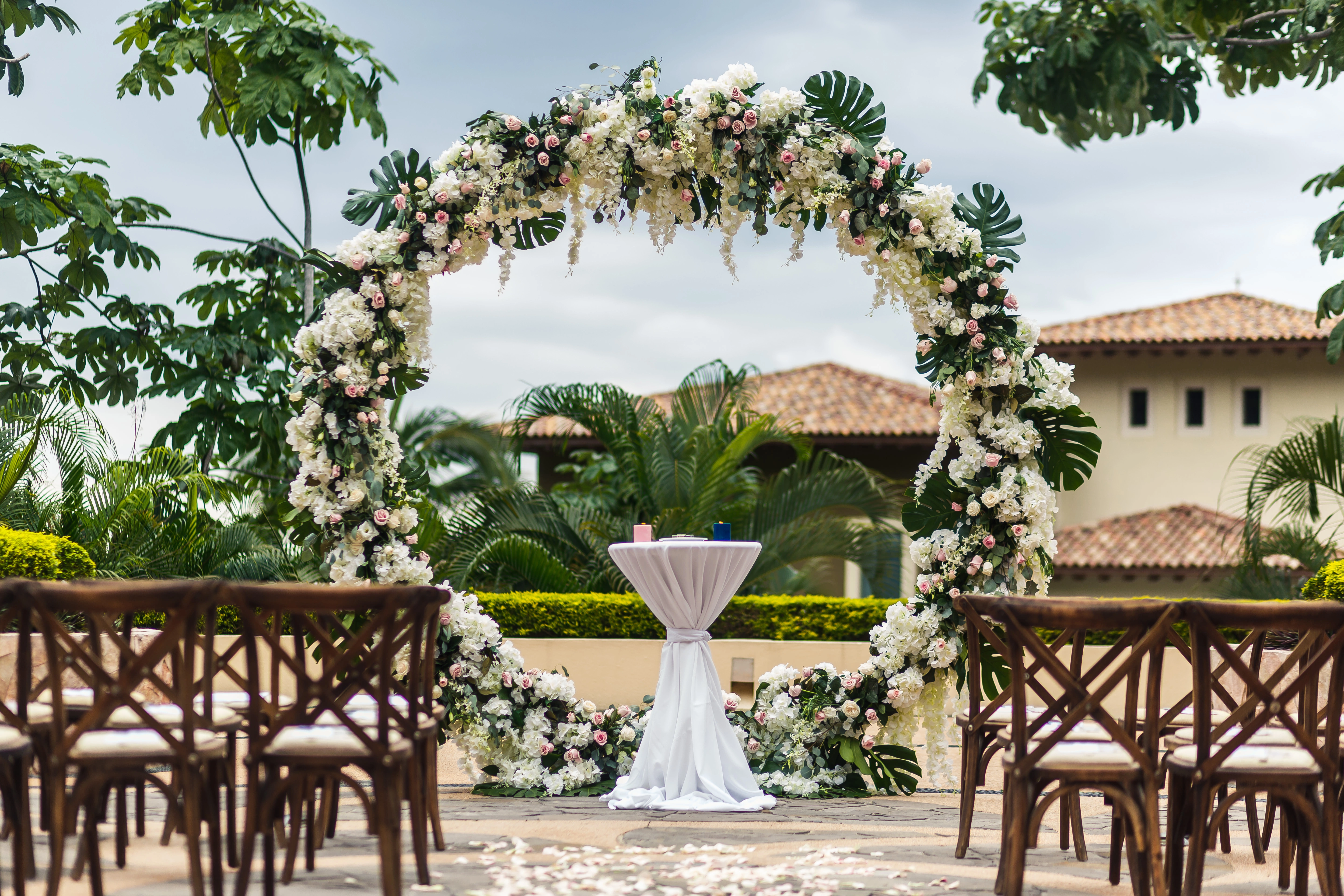 Floral arch and elegant chairs set up for an outdoor wedding ceremony, with tropical greenery and a villa in the background.