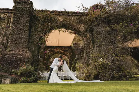 The groom dips and kisses the bride, in a long veil, on the lawn in front of ivy-covered stone arches