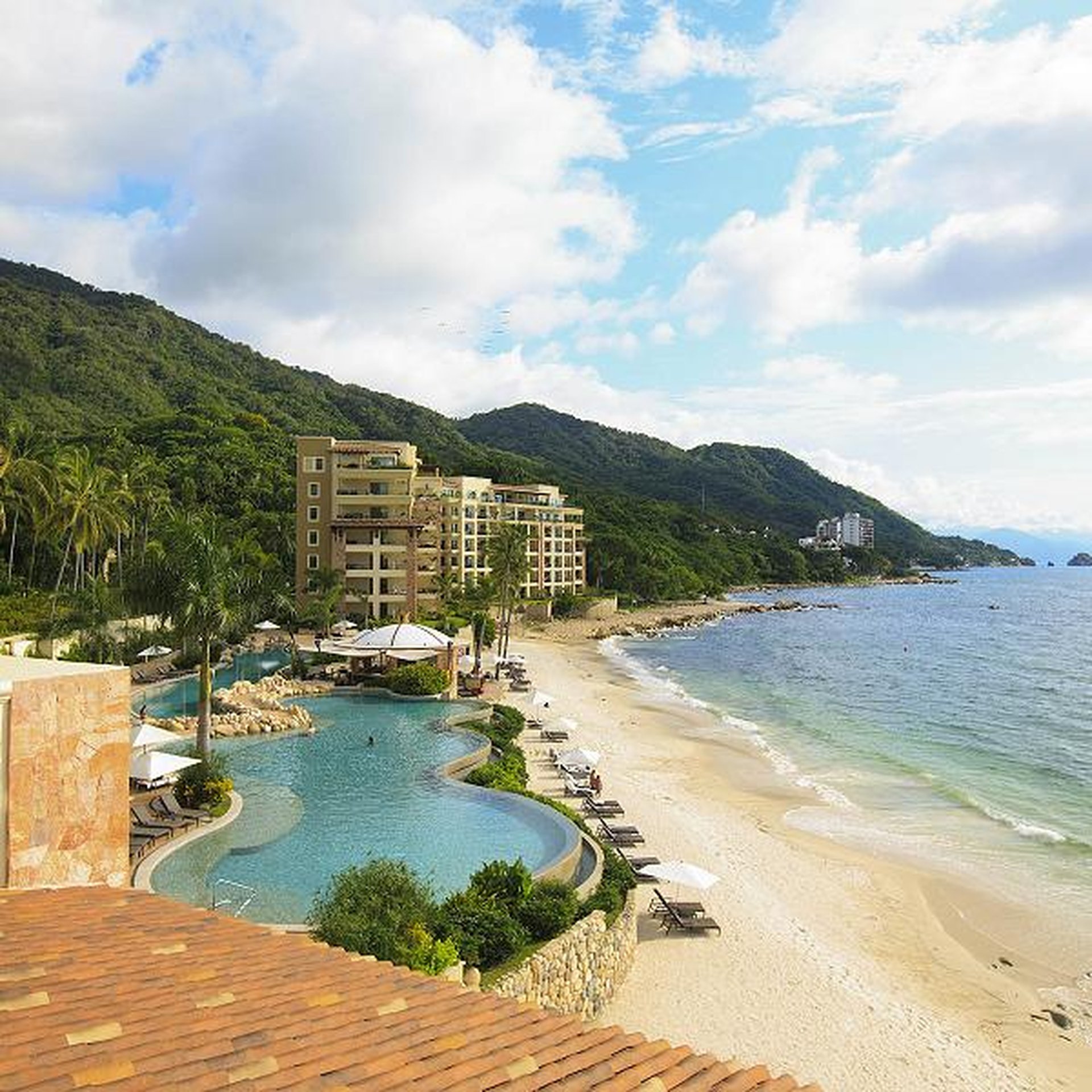 Beachfront resort with a curved pool, sun loungers, and umbrellas, surrounded by lush green hills and overlooking a calm ocean under a partly cloudy sky.