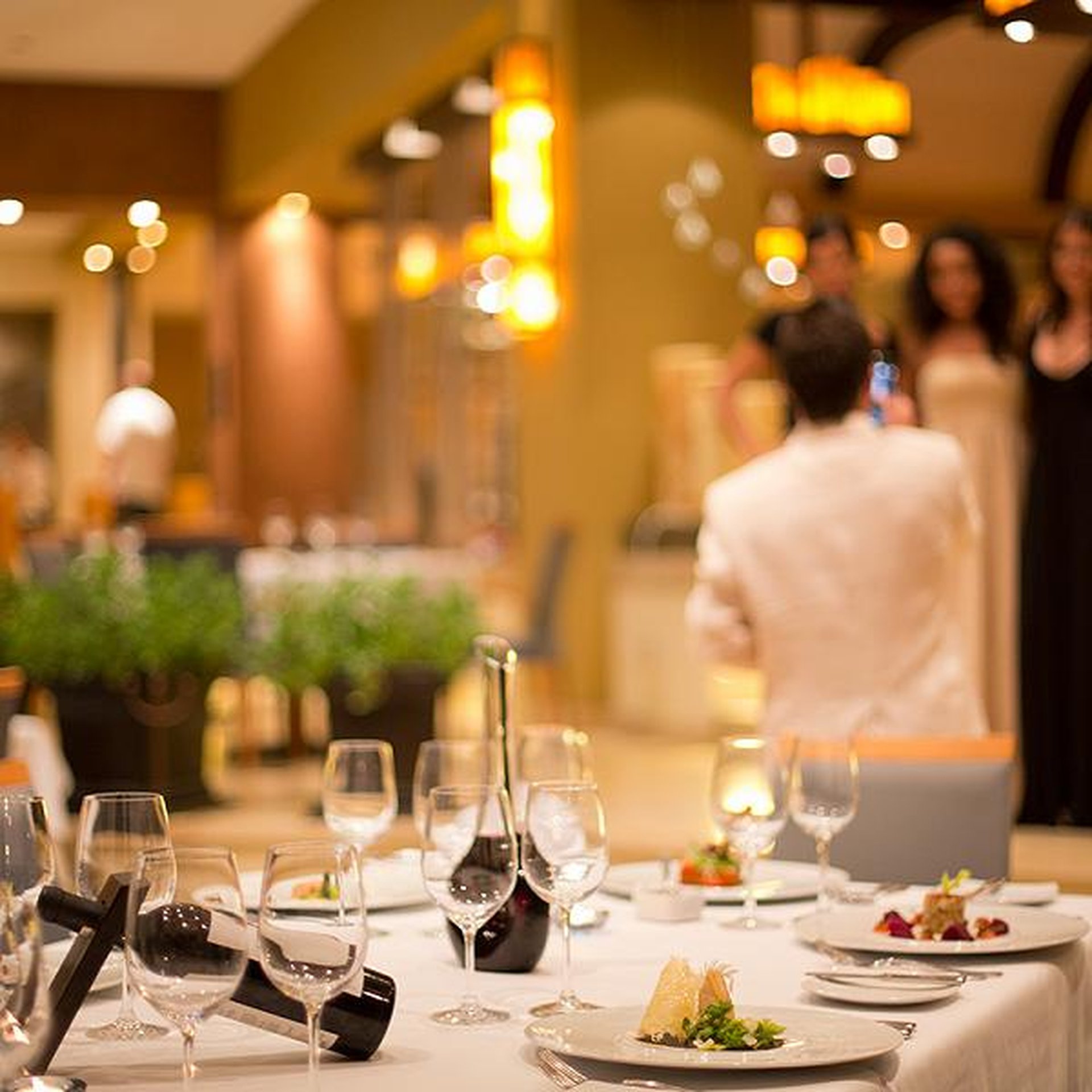 Elegant restaurant setting with a table set for dining, featuring wine bottles, glasses, and plates. A person in white takes a photo of a group.