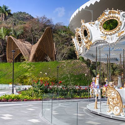 A white and gold carousel with animal figures is set near lush greenery and a unique bamboo structure under a clear blue sky.