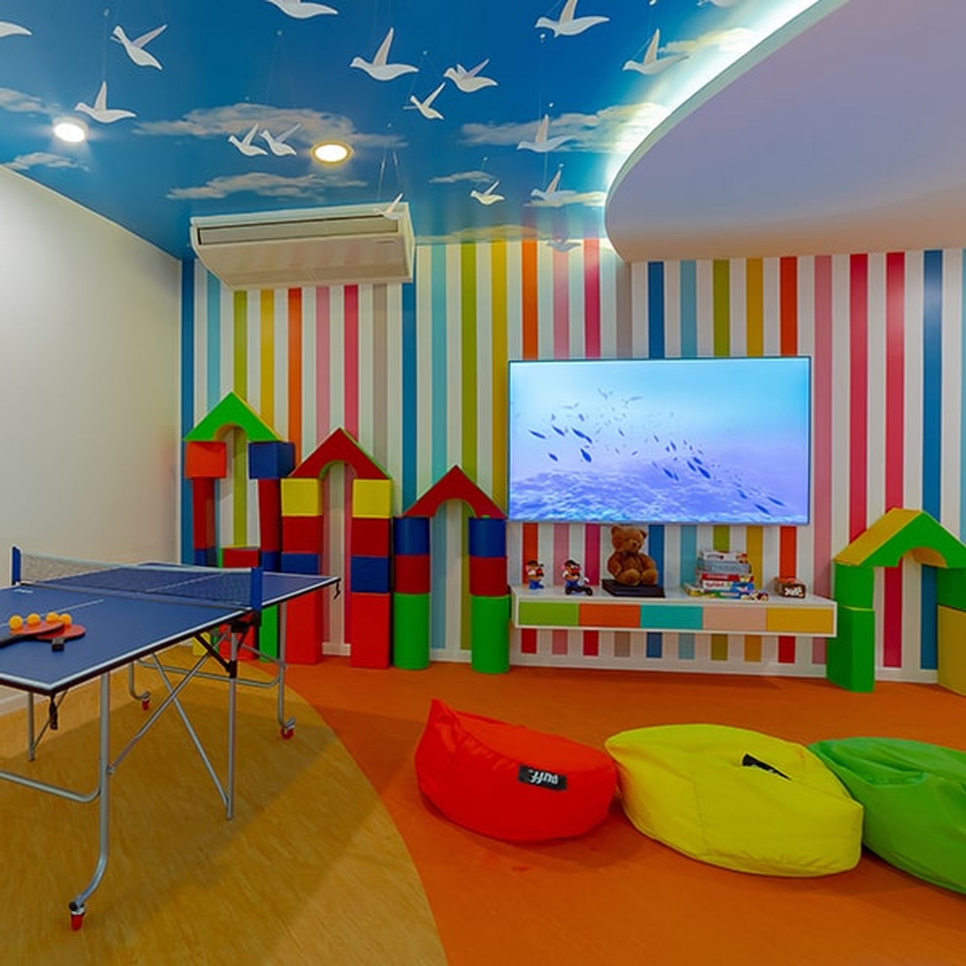 A colorful playroom with a striped wall, bean bags, a TV, toy blocks, and a ping pong table under a ceiling with bird decals.