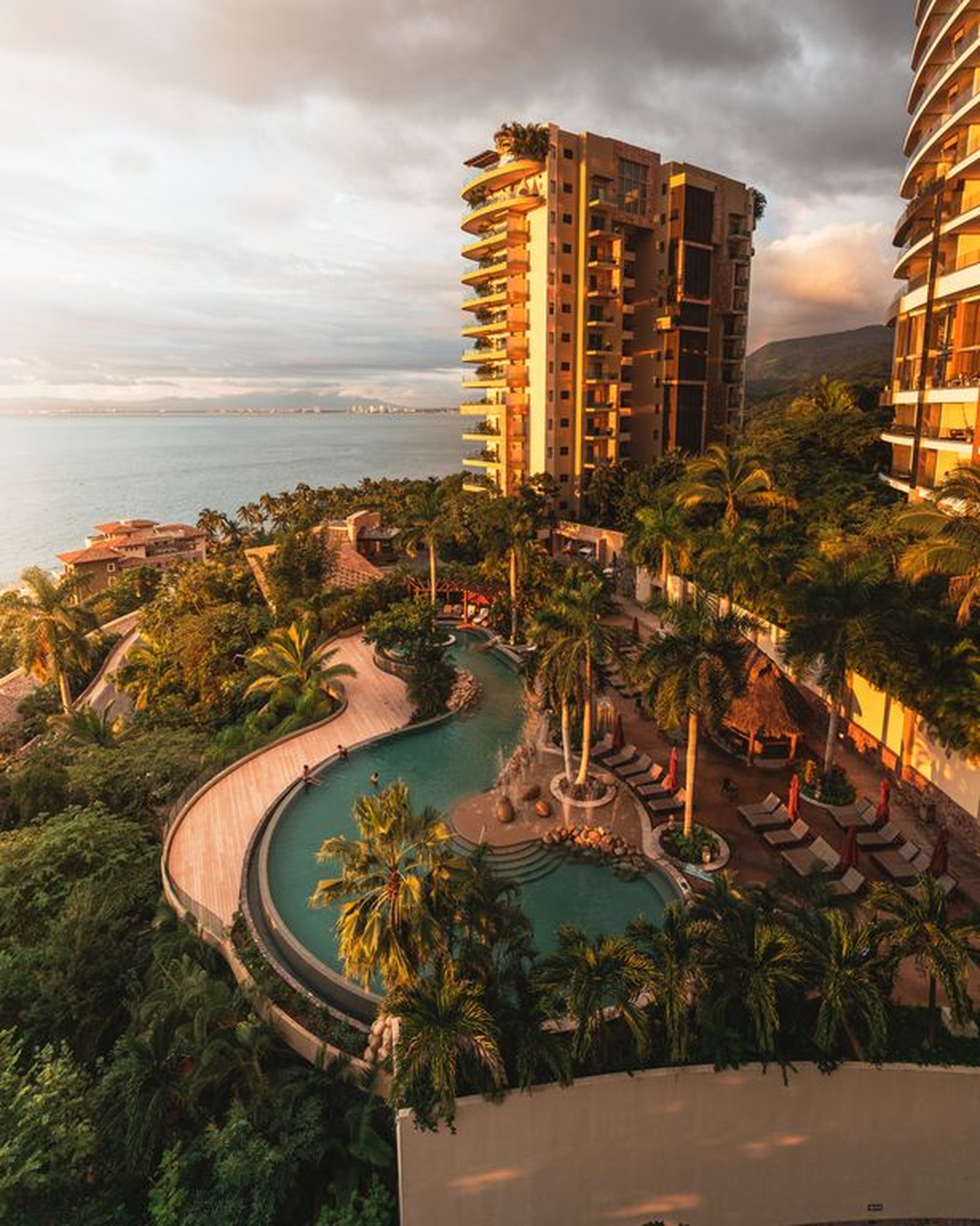 Aerial view of a luxury resort with a circular pool, surrounded by lush greenery and tall buildings, overlooking the ocean at sunset.