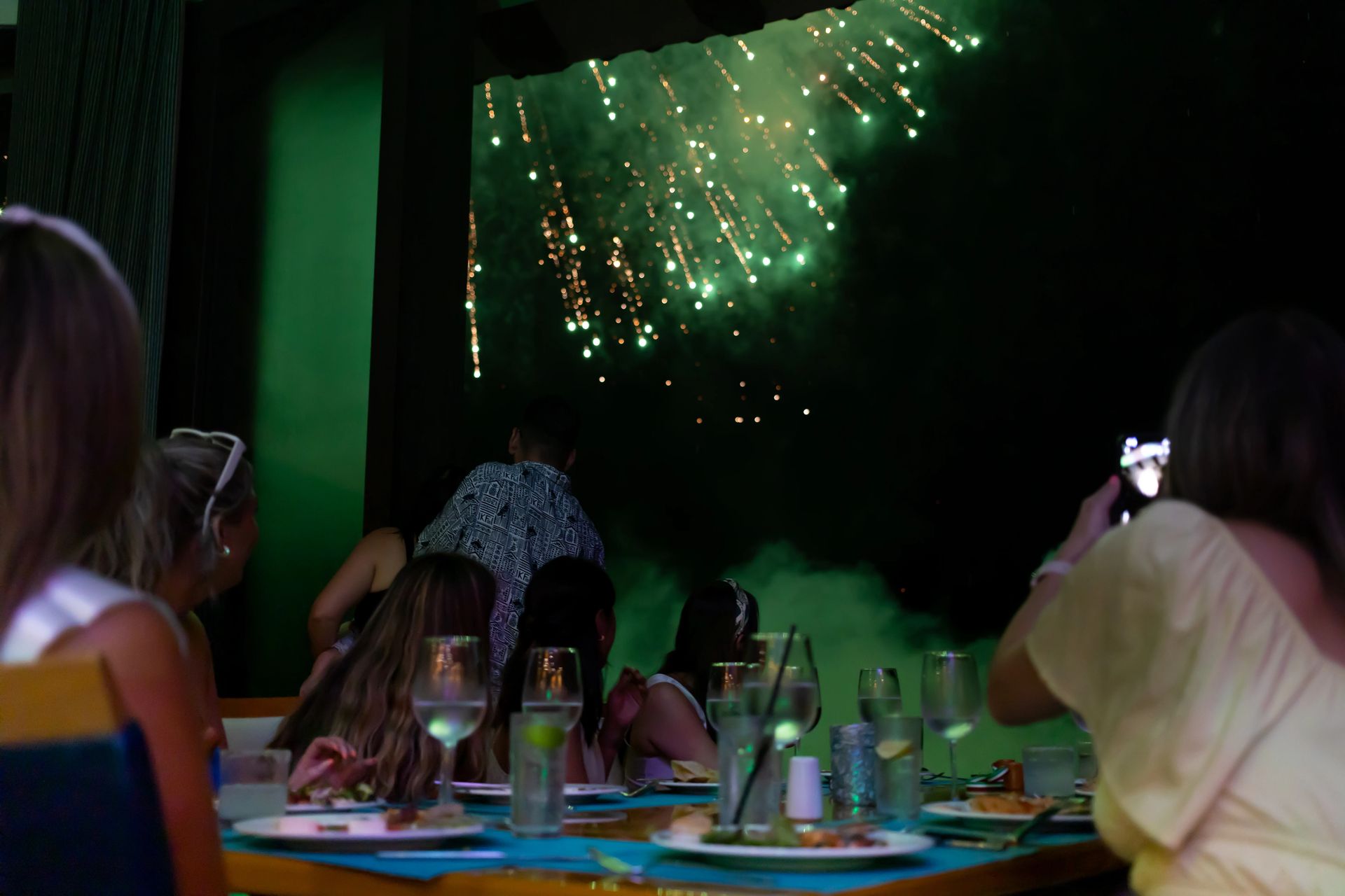 Guests enjoying a firework display over a dark sky, seated at a table with drinks and food, creating a festive atmosphere.