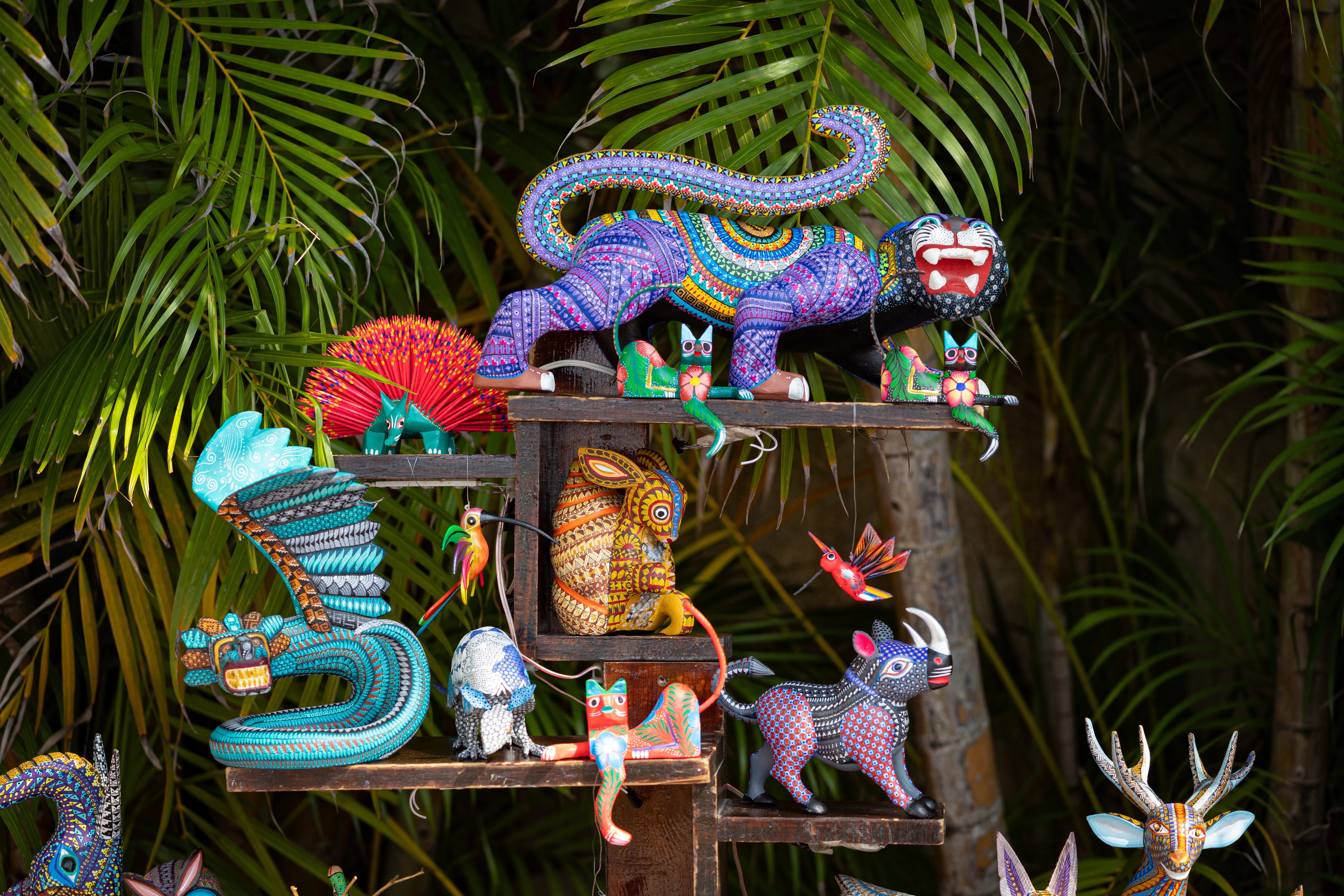 Colorful Mexican folk art sculptures, including jaguars, birds, and mythical creatures, displayed against lush tropical foliage.