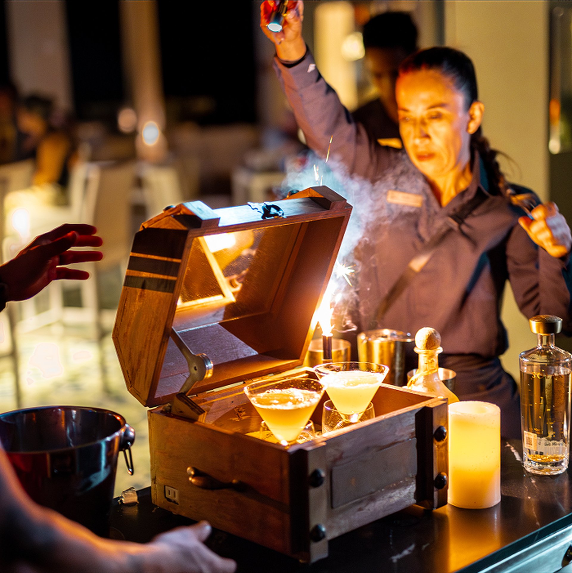 A bartender performs a flaming cocktail presentation with two martini glasses in a wooden box, surrounded by bottles and a lit candle.