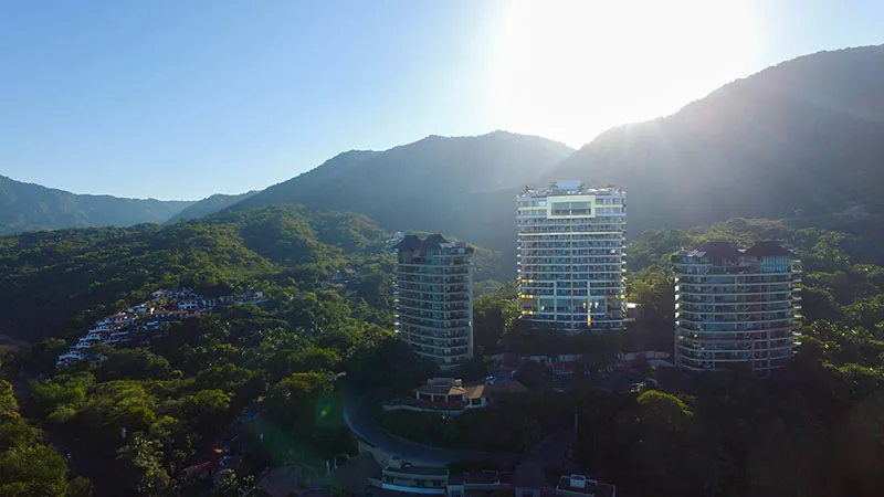 Aerial view of a resort surrounded by lush green hills and mountains under a bright sunlit sky.