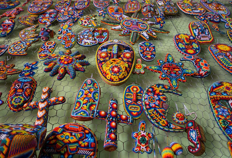 Colorful Huichol beaded art displayed on a mesh background, featuring masks and various shapes with intricate patterns and vivid designs.