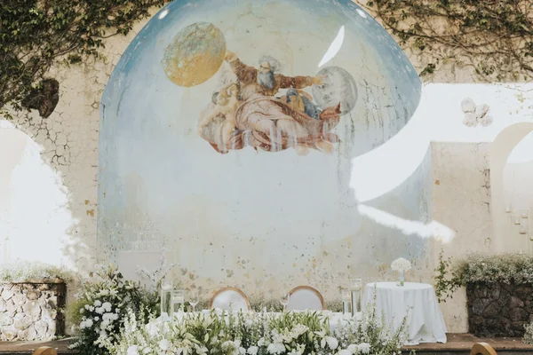Mesa larga de banquete con sillas de respaldo redondo y flores blancas junto al muro de hiedra