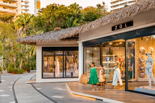 Woman and two girls exit ZOL boutique at Garza Blanca Puerto Vallarta with shopping bags beneath a palm-thatched roof at sunset.