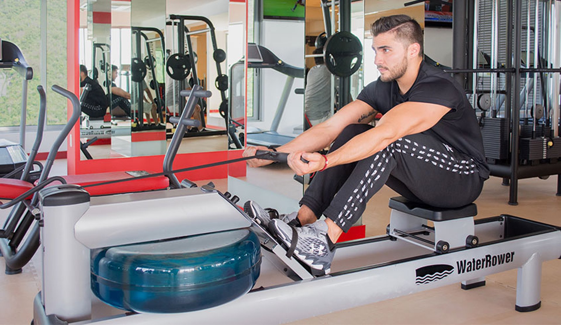 Man exercising on a WaterRower machine in a gym, wearing a black shirt and patterned pants. Fitness equipment is visible in the background.