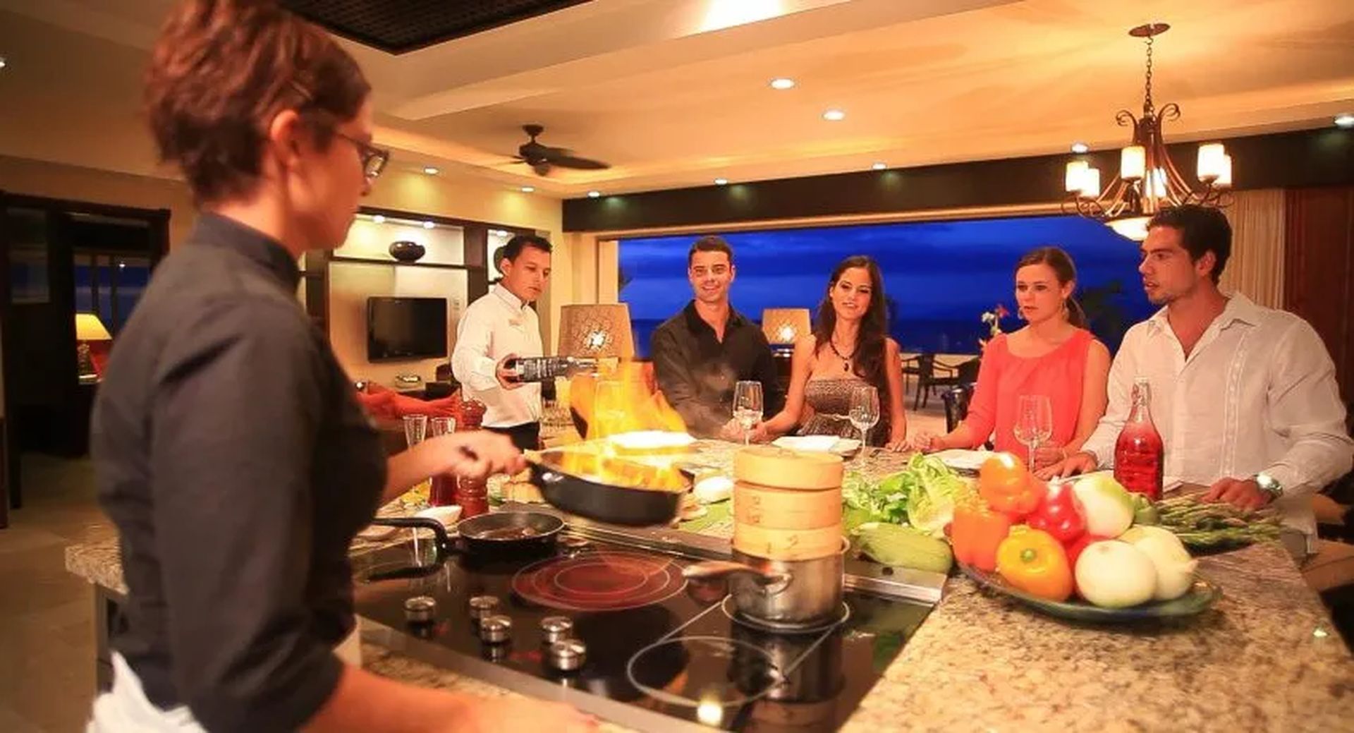 A chef flambés a dish in a stylish kitchen while five guests watch, enjoying wine and conversation at an upscale Cancun resort.