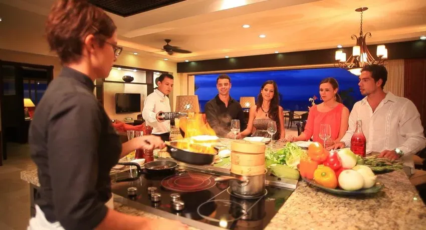 A chef flambés a dish in a stylish kitchen while five guests watch, enjoying wine and conversation at an upscale Cancun resort.