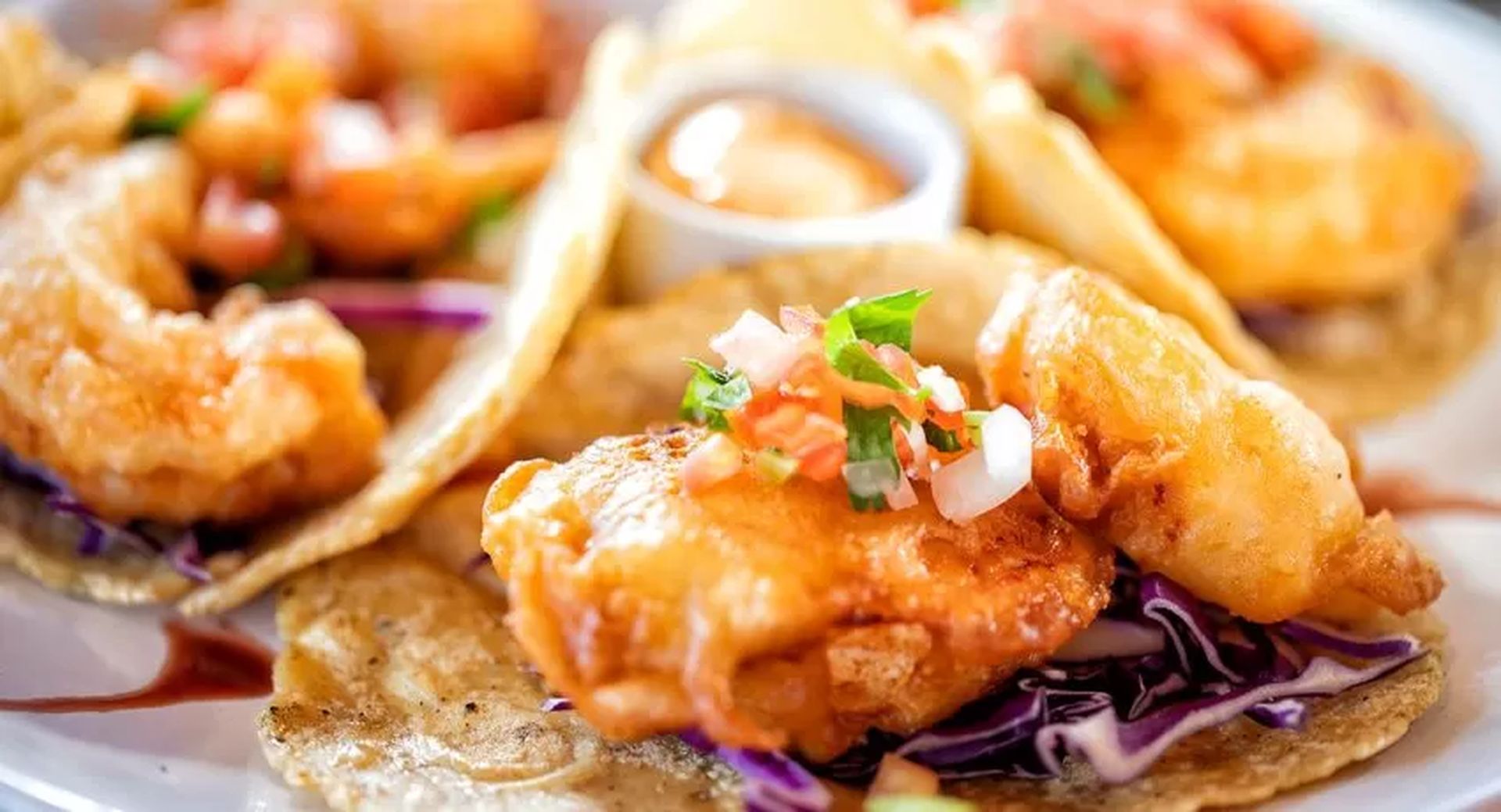 Close-up of crispy fried fish tacos with cabbage slaw, topped with diced tomatoes and a side of dipping sauce.