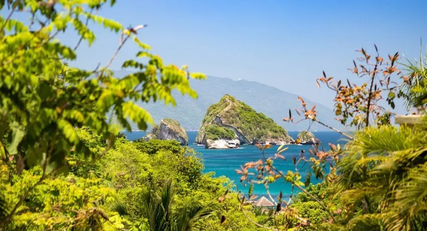 Scenic view of lush greenery framing rock formations in turquoise ocean, under a clear blue sky.