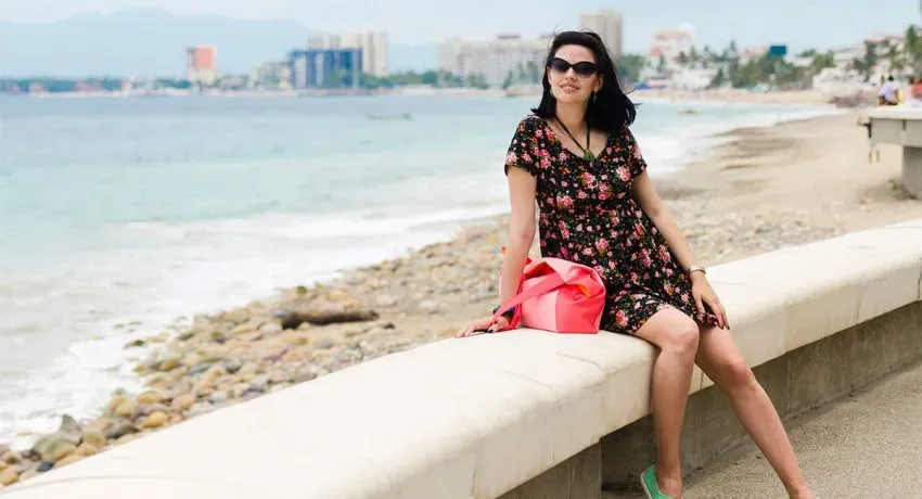 Woman in a floral dress sits on a beachside wall, with a cityscape and ocean in the background on a sunny day.