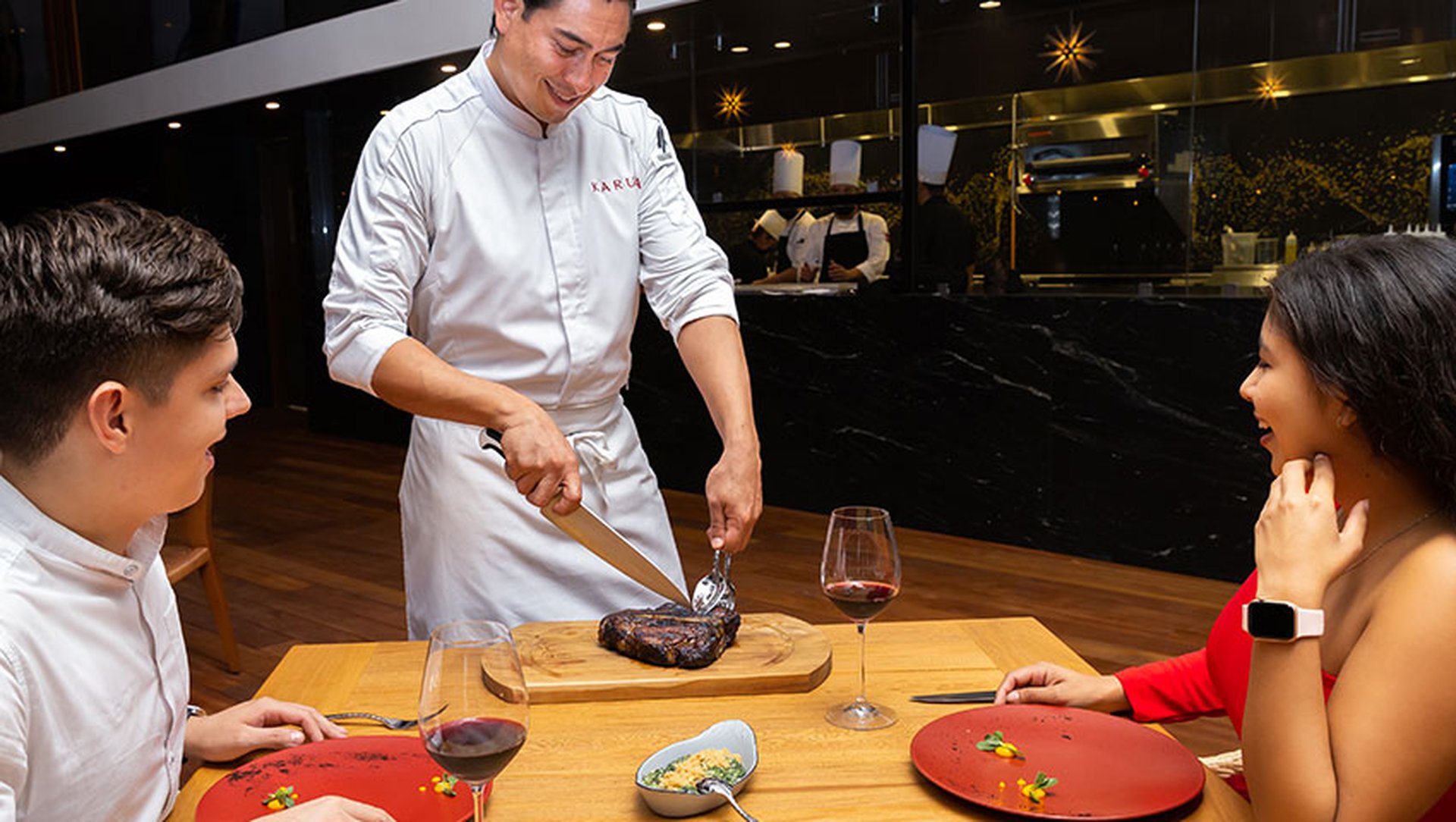 A chef slices a steak at a table for a smiling couple seated in a restaurant, with glasses of red wine and plates in front of them.