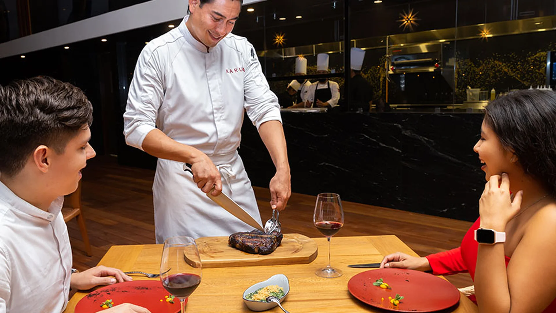A chef slices a steak at a table for a smiling couple seated in a restaurant, with glasses of red wine and plates in front of them.