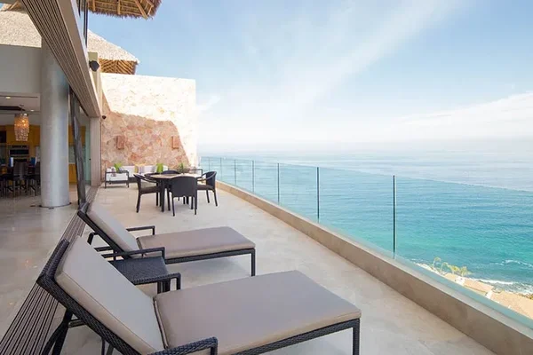 A luxurious oceanfront balcony with lounge chairs, a dining table, and glass railing, overlooking a vast, calm sea under a clear sky.