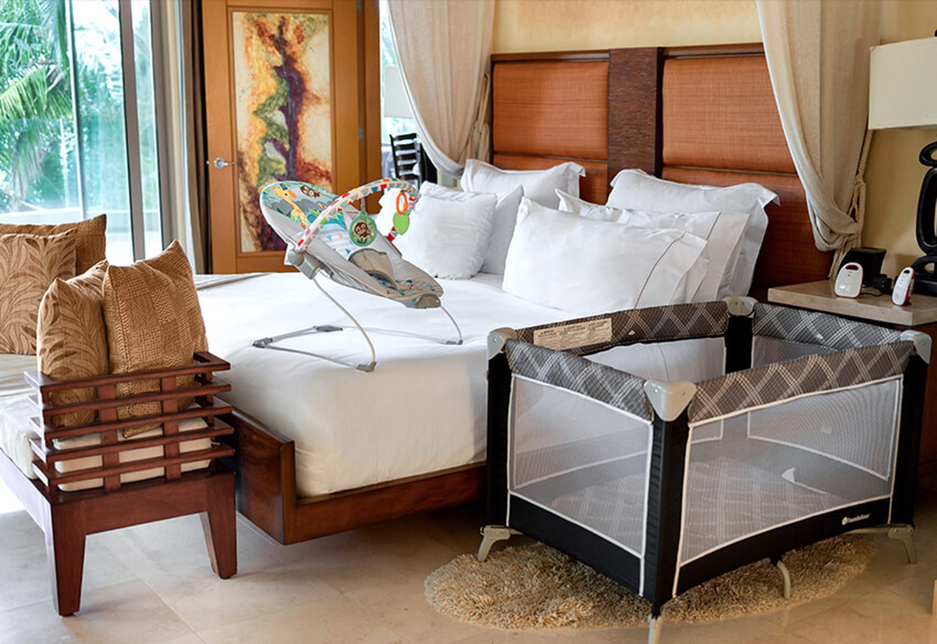 Garza Blanca luxury suite: king bed with white linens, wooden headboard, drapes; a baby jumper on the bed beside a portable play yard.