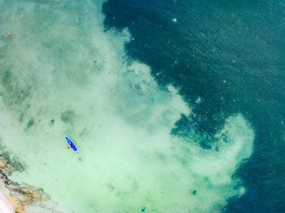 Aerial view of a paddleboarder on clear turquoise waters near a sandy shore, surrounded by deeper blue ocean.