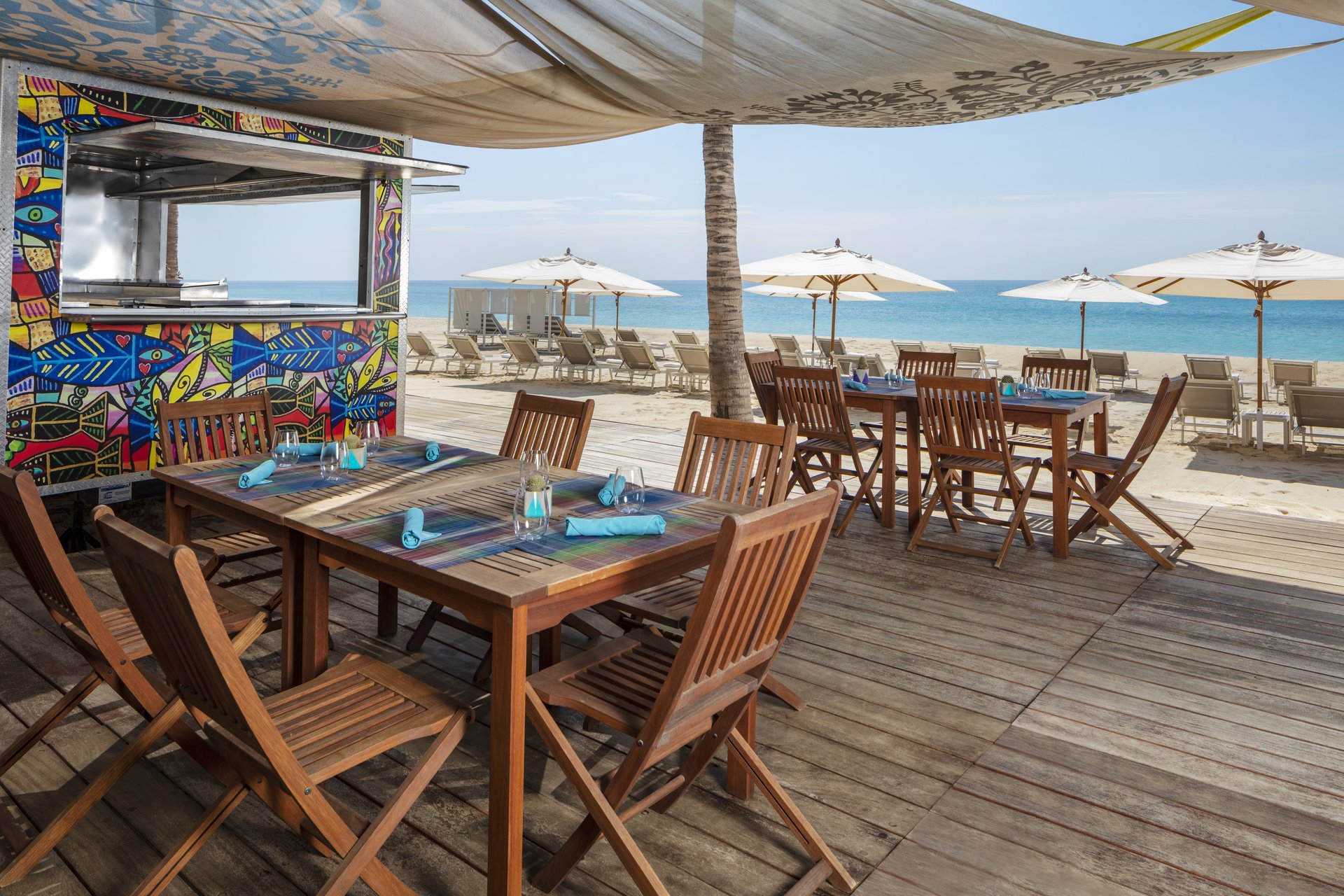 Beachside seating area with white wooden chairs and tables on a wooden deck, shaded by fabric canopies. Umbrellas and the sea are in the background.