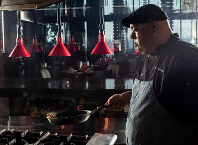 Chef in a dimly lit kitchen cooking with a frying pan, wearing a black cap and apron, with red pendant lights in the background.