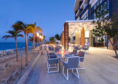 Outdoor restaurant with modern seating, ambient lighting, and ocean view at dusk. Palm trees and decorative fire elements enhance the atmosphere.