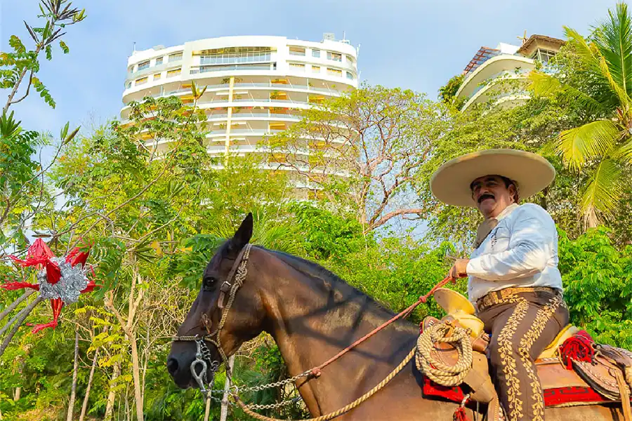 A man in traditional attire on horseback beside tropical foliage and a tall, curved modern building with a clear blue sky above.