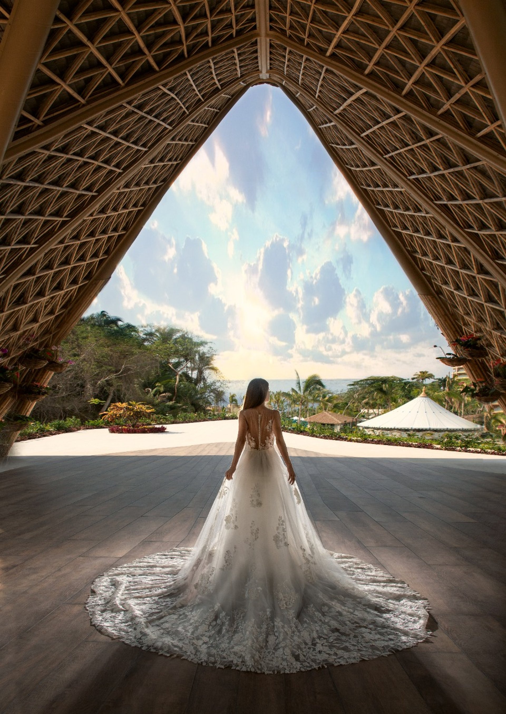 Bride in an elegant gown stands under a grand wooden arch, gazing at a tropical garden and ocean view under a vibrant sky.