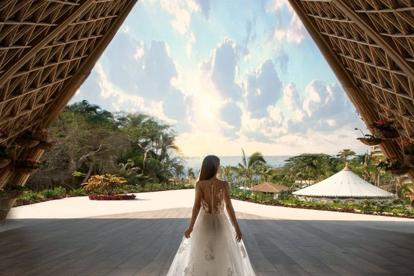 Bride in an elegant gown stands under a grand wooden arch, gazing at a tropical garden and ocean view under a vibrant sky.
