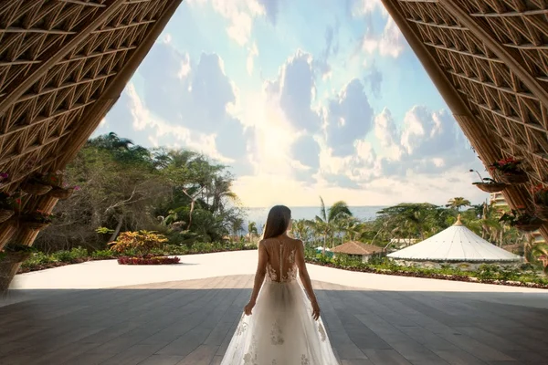 Bride in an elegant gown stands under a grand wooden arch, gazing at a tropical garden and ocean view under a vibrant sky.