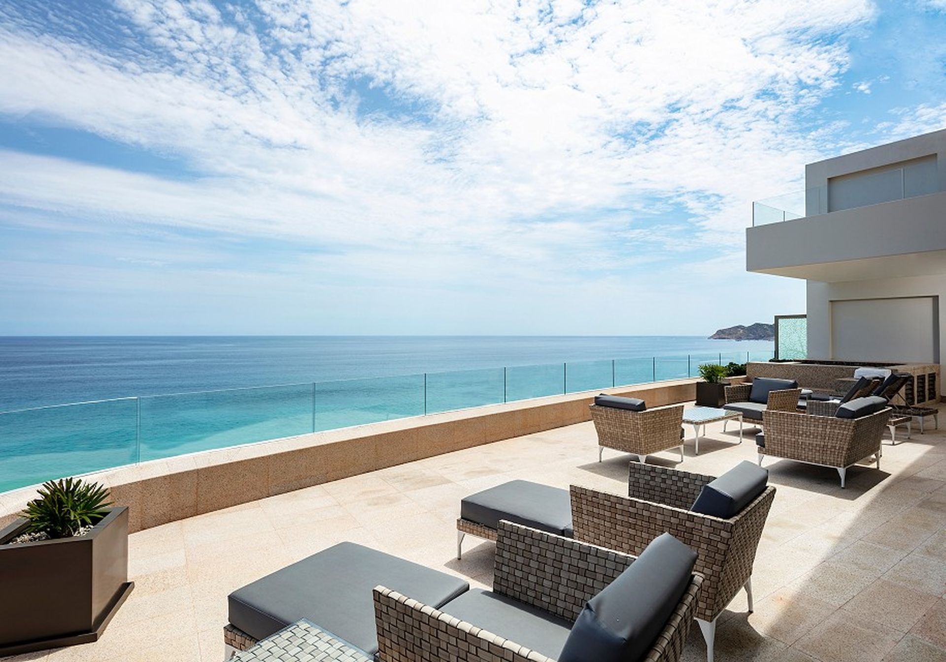 Modern terrace with wicker lounge chairs overlooking a calm ocean under a partly cloudy sky. Glass railing enhances the view.