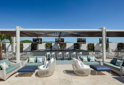 Rooftop lounge with modern seating, a bar with stools, multiple TVs, and a pergola, set against a clear blue sky.