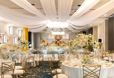 Elegant banquet hall with white draped ceiling, round tables adorned with floral centerpieces, and gold chairs on a patterned blue carpet.