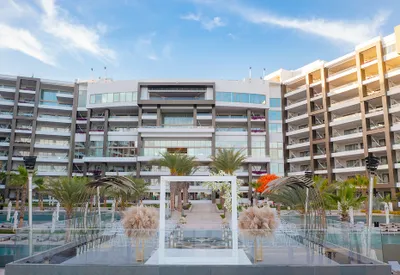 Modern resort with palm trees, a pool, and a decorative archway, set against a backdrop of a multi-story building and a blue sky.