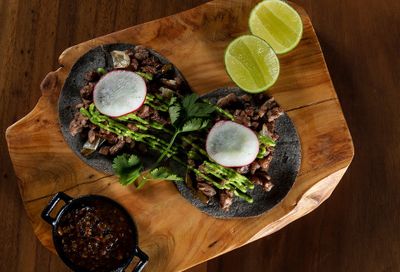 Two tacos with meat, avocado sauce, and radish slices on a wooden board, garnished with cilantro and lime halves, next to a small bowl of salsa.