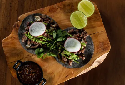 Two tacos with meat, avocado sauce, and radish slices on a wooden board, garnished with cilantro and lime halves, next to a small bowl of salsa.
