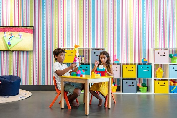 Two children play with colorful blocks at a small table in a vibrant playroom with striped walls and organized storage cubes.