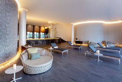 Modern spa lounge with wicker chairs, loungers, soft lighting, and elegant decor. Walls feature floral patterns, creating a serene atmosphere.