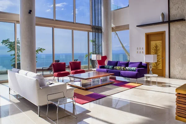 Modern living room with ocean view, featuring colorful sofas, red chairs, a glass coffee table, and tall windows letting in natural light.